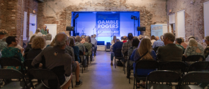 Audience seated in a rustic brick-walled venue facing a stage with "Gamble Rogers Concert Series" banner and musical equipment.