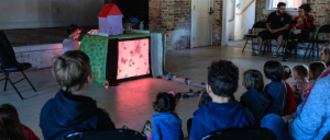Children and adults watch a puppet show with a house-shaped stage and illuminated screen in an indoor brick-walled room.