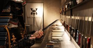 Samurai armor holding a Japanese kitchen knife inside a MUSASHI JAPAN store with knives displayed on the wall.