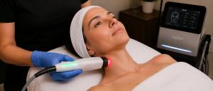 Medspa esthetician wearing blue gloves uses a handheld XERF device to perform a light-based skin treatment on a female client's neck, with the XERF diagnostic screen visible in the background.