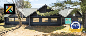 Black wooden cabin with teal door and large windows, surrounded by trees and grass under a clear sky.