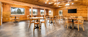 Interior of a spacious wooden cabin dining area with round tables, wooden chairs, large windows, and a mounted flat-screen TV.