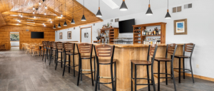 Spacious bar area with wooden counters, high stools, pendant lighting, and additional seating along the back wall under a wood-paneled ceiling.