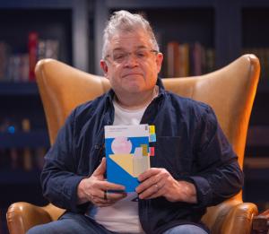 Patton Oswalt poses in a high-backed leather chair, holding a copy of "This Paradise" by Ruby Cowling.