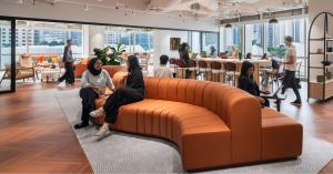 Diverse professionals working and conversing inside a Tailored by Flexi modern flexible workspace with a large curved orange sofa, herringbone wood flooring, and floor-to-ceiling windows showing city skyscrapers.