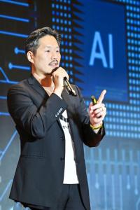 David Johnson Jiang speaking on stage with a microphone, gesturing with one hand in front of a large screen displaying the letters “AI” and digital graphics.