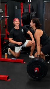 Train With Dave personal trainer kneeling beside and encouraging a smiling female client as she performs a loaded trap-bar deadlift in an Orange County gym.