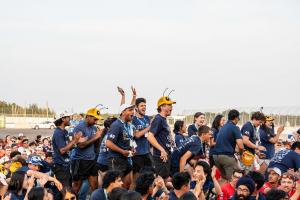 A group of students in navy team shirts celebrate outdoors at a racetrack, several wearing yellow-and-black bee-antenna headbands and event medals. One student claps, others cheer with open mouths, surrounded by a large crowd.