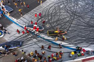 Aerial view of a speedway paved surface covered in dark, swirling tire marks. Teams in red and yellow crew shirts carry low, solar-panel-topped vehicles across the track by hand, surrounded by clusters of spectators and support crew.