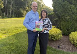 A smiling middle-aged man with short white hair and glasses, wearing a light blue checkered button-down shirt and dark pants, stands outdoors on a sunny grassy lawn beside a smiling woman with dark shoulder-length hair, large hoop earrings, a black-and-wh