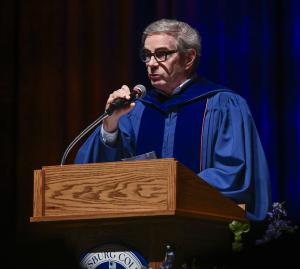 James G. Basker, wearing blue academic regalia, speaks from a podium during the 2026 Master of Arts in American History commencement.