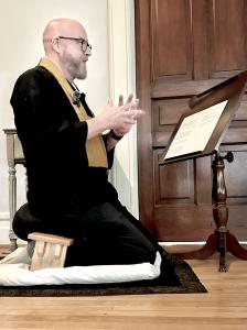 Sensei Michael Brunner of Ottawa, Illinois, wearing traditional Zen robes and a lapel microphone, kneeling on a wooden meditation bench while delivering a lecture from a stand.