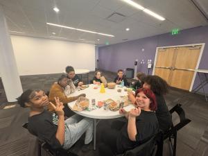 Seven middle school students and a mentor seated around a table sharing a meal together, smiling at the camera.