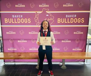 Smiling middle school student wearing a vest and tie holding a certificate, standing in front of a maroon and gold "Baker Bulldogs" backdrop.