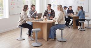 A group of college students collaborating around wooden tables in a campus classroom while sitting on ergonomic standing desk stools with circular non-slip wobble bases.