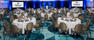 Banquet hall at Hilton Phoenix/Mesa set with round tables, chairs, white linens, folded napkins, and two large screens.