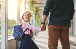 A school aged girl with glasses, braids and a big smile holds her parent's hand and looks back at camera.