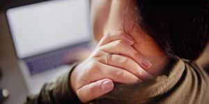 Woman holding the back of her neck while using a laptop, representing tech neck caused by prolonged screen use.