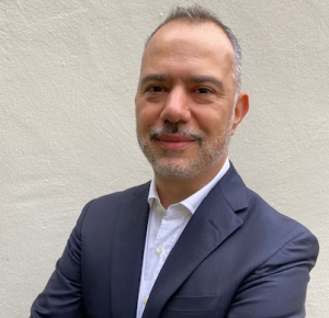 Portrait of a man with short gray hair and a trimmed beard wearing a dark suit jacket and white dress shirt, smiling with his arms crossed against a light-colored wall.