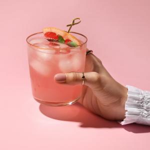 A paloma cocktail in a tall glass with a grapefruit garnish, held against a pink background.