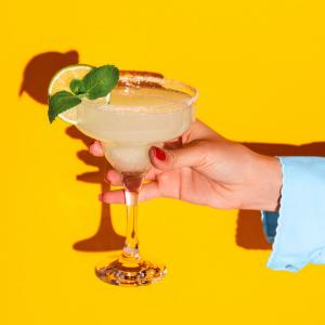margarita in a stemmed glass with a lime wheel and mint garnish, held against a yellow background.