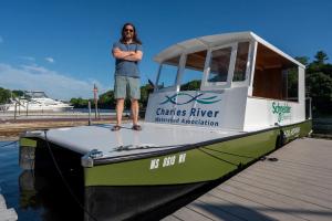 Drew Rollert stands with arms crossed on the deck of Just Get On, Wada Hoppah's green-and-white electric ferry, docked on the Charles River.