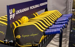 Black and yellow Freedom Fighter Paddles with blue grips displayed on a Ukrainian Pickleball Federation rack, inscribed with the Shevchenko line "Борітеся – поборете!" alongside a yellow pickleball.