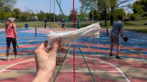A hand holds the transparent Anywhere Protractor template and white measuring cord in front of an outdoor multi-use court where two players are pickling in the background, demonstrating how quickly a regulation court can be set up anywhere.