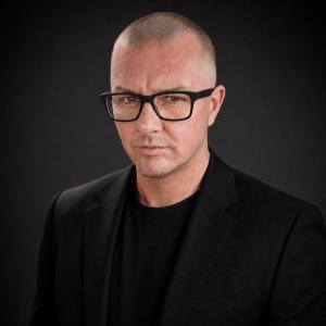 Professional studio headshot of author and technology entrepreneur Jason Ansell wearing a black blazer against a dark background.