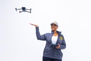 Steffanie Rivers wearing a TCB Drones jacket and cap holds a drone controller beneath a flying drone against a white background.