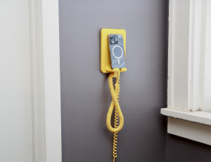 Coilie, the modern landline: a rounded sunshine yellow wall-mounted phone perch on a slate gray wall beside white trim, holding a docked smartphone, with a retro yellow coiled charging cord spiraling down toward the floor.