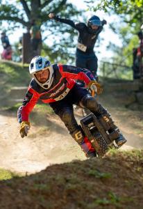 An electric unicycle racer wearing a red jersey leans into a berm on an off-road dual slalom course