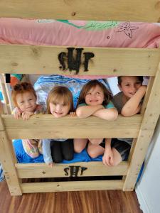 Four young children smiling and peeking over the rails of a wooden bunk bed marked with the SHP logo, with pink and blue bedding visible.