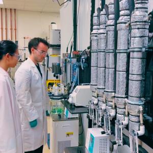 Two researchers in lab coats observe Precision Periodic's 1/10th scale Multi-Scale Refining Unit in a laboratory, featuring vertical cylindrical reactor columns used for battery-grade nickel processing.
