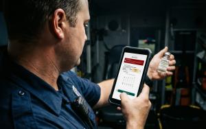 Paramedic holding a medication vial and a rugged mobile device displaying the OneDose eMACC™ medication verification screen.
