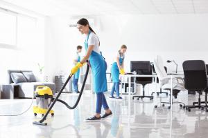 Three cleaning staff in blue aprons and yellow gloves servicing an open plan office, one vacuuming the tile floor while two wipe down desks and monitors