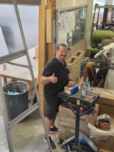 A man smiles and gives thumbs up. There are LiTime batteries visible and they are in a warehouse working on a boat with tools in the background