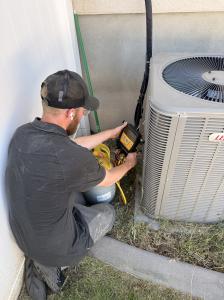 An HVAC technician kneeling next to a residential outdoor air conditioner, using a yellow refrigerant cylinder and digital gauges to service the HVAC system.