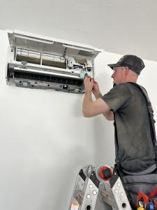 A professional HVAC technician standing on a ladder, installing the internal components of a white wall-mounted ductless air conditioning unit.