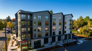 Daytime elevated view of the Cartwright Apartments, a five-story building with white and gray siding, black balconies and ground-floor storefronts, beside a residential street under a clear blue sky.