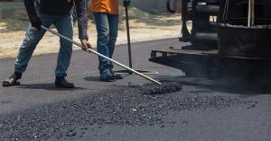 Two paving crew members in jeans, long sleeves and work gloves using a shovel and a long rake to spread a pile of fresh black asphalt at the rear of a commercial paving machine, with steam rising from the hot mix.