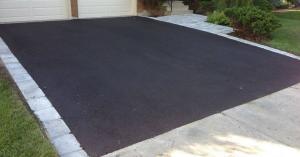 Newly paved black asphalt residential driveway bordered on three sides by light gray cobblestone paver stones, leading up to a two-car beige garage with a paver-stone walkway and landscaping along the right side.