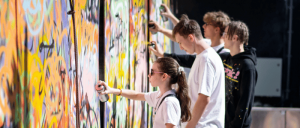 Teens spray painting colorful graffiti art on a large outdoor wall in bright daylight.