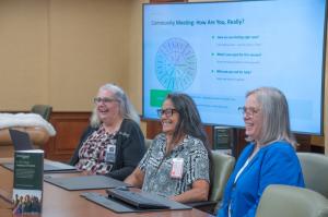 Three female OneQuest Health staff members, wearing professional attire and ID badges, sit at a large wooden boardroom table, all smiling and laughing genuinely while looking off-camera to the right. In the foreground is a small branded brochure titled "O