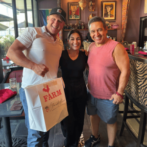 Three smiling people posing indoors, one holding a "FARM and Clandestino" paper bag, with colorful wall art and eclectic decor in the background.