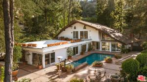 A two-story white stucco A-frame house with a shingle roof and a large flat deck is nestled on a forested hillside. Below the house, a swimming pool is surrounded by a large paved patio with various potted plants.
