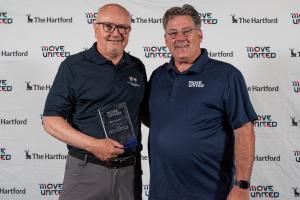 Mike Mushett and Glenn Merry hold an award in front of a Move United backdrop