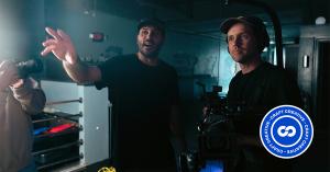 Two video production crew members direct a scene on set, with one operating a professional cinema camera.