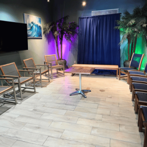 Modern waiting room with wooden chairs, tropical plants, a large TV, and a blue curtain backdrop under colorful lighting.