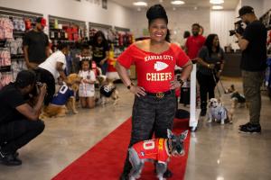 Audrey Morrow stands with Buddy on a red carpet inside a dog-apparel store while photographers, families and dogs in sports jerseys participate in a community casting-call scene.